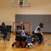 Opposing adult teams reaching for the ball with hockey sticks during a wheelchair floor hockey game.